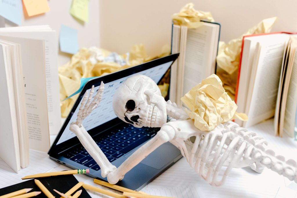 Skeleton-with-skull-in-laptop-screen-surrounded-by-pencils-books-and-screwed-up-paper