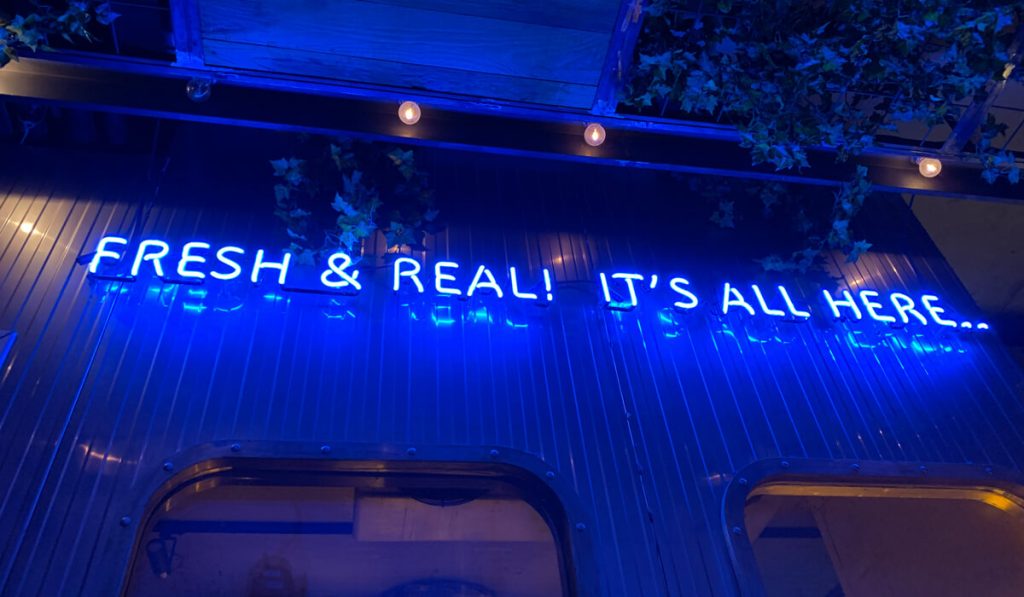 Blue-neon-sign-reads-Fresh-&-Real!-It’s-All-Here